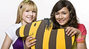 girl wearing pink cap-sleeved shirt holding yellow and black stripe cut-out animal standee beside girl wearing purple and white cap-sleeved shirt