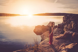 woman in black and red dress standing on a rock near body of water during golden hour HD wallpaper