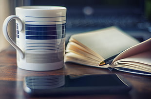 white and blue ceramic mug near brown notebook and black pen on brown desk