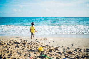 toddler in yellow shirt and gray shorts standing at seashore near blue sea during daytime HD wallpaper