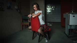woman wearing white and black cap-sleeved shirt and red, black, and white plaid skirt sitting on red padded chair near white painted wall in room