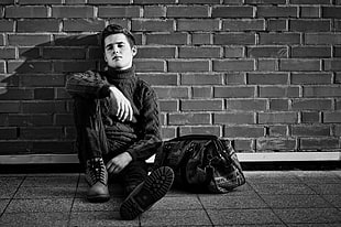 grayscale photography of man sitting and leaning on brick wall with duffle bag HD wallpaper