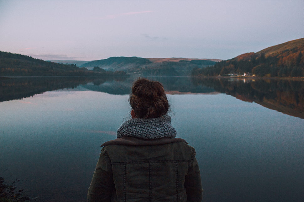 person wearing gray jacket in front of  body of water and mountain HD wallpaper