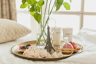grey metal Eiffel Tower beside two disposable cups, fragrance bottle, cupcakes, and glass vase place on round brass-colored tray near window at daytime HD wallpaper