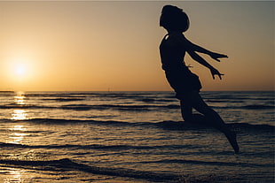 silhouette of woman jumping near ocean water during yellow sunset HD wallpaper