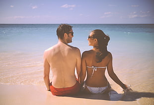 couple sitting beside each other on sand beside ocean during daytime HD wallpaper
