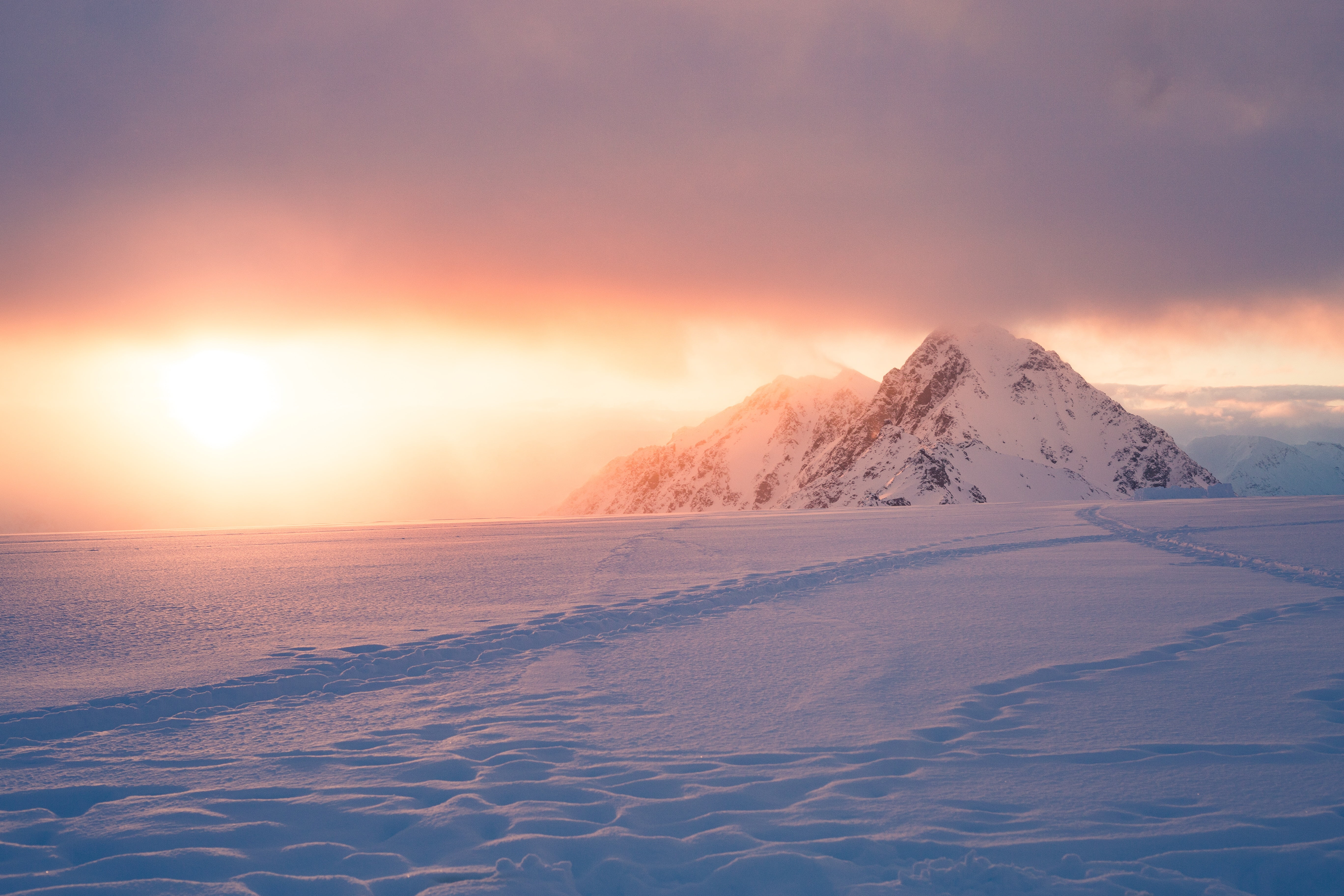 snow covered mountain and ground with trail tracks under cloudy sky