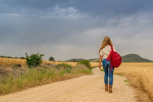 woman wearing white top and blue denim jeans with red backpack walking on rocky road near brown grass field at daytime HD wallpaper