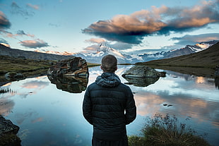 man facing the water with rocks under the gray cloudy sky