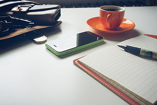 green iPhone 5C near orange ceramic teacup with saucer beside white lined book with pen