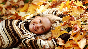 close up photo of woman wearing beige and brown striped sweater lying on bed of maple leaves
