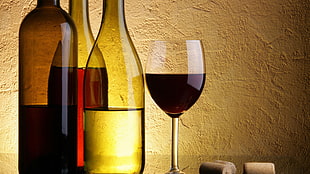 half filled clear wine glass beside three clear glass wine bottles and three corks