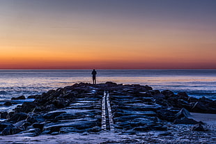 silhouette photo of person standing on rock facing sea during sunset