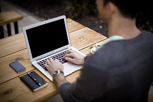 depth of field photography of man using MacBook Air on brown wooden table beside book and black iPhone 4