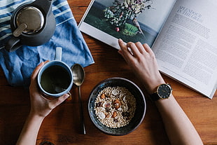 person holding blue ceramic mug while opening a book