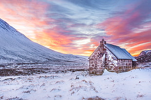 Brown House Surrounded by Snow Covered Field Near Snow Covered Mountain Under Yellow Blue and Orange Sunset HD wallpaper