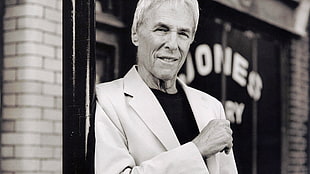 grayscale photo of man wearing suit jacket leaning on black metal pole