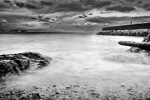 grayscale photo of river and shore stones with dock under cumulus clouds