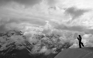 grayscale photo of a person standing on cliff carrying snowboard near snow covered mountain surrounded by clouds