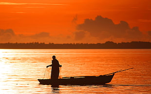 silhouette photo of person holding fish net standing on canoe on body of water under orange sky HD wallpaper