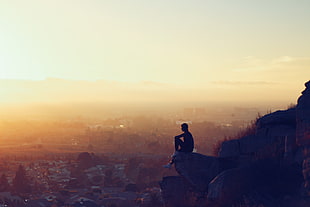 landscape silhouette photography of person sitting on cliff facing urban area during golden hour HD wallpaper