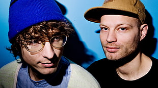 man in brown cap and black shirt beside man in black knit cap, eyeglasses, and blue shirt