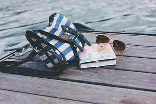 blue, white, and black stripe crossbody bag beside silver-framed aviator style sunglasses and book on brown wooden dock HD wallpaper