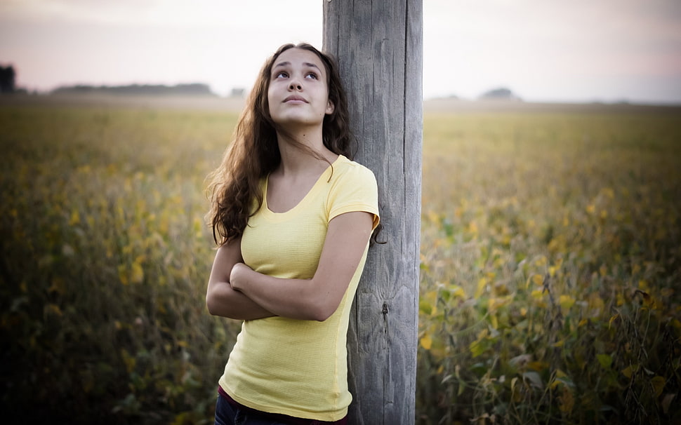 woman in yellow scoop-neck shirt leaning on brown wooden pole beside a white and yellow flower field HD wallpaper