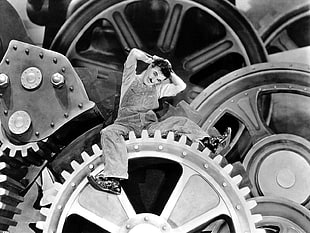 grayscale photo of man in overalls sitting on gear