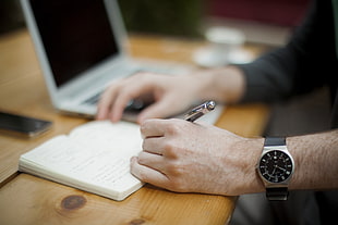 man wearing silver analog watch holding a pen writing on a book