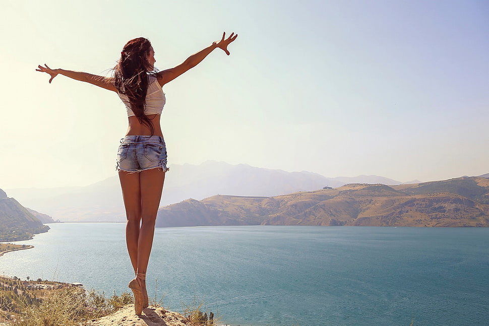 woman in white crop top and micro denim shorts standing on the hill during daytime HD wallpaper