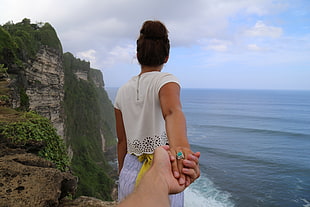 woman wearing white shirt and purple bottoms holding person's hand standing on cliff near body of water