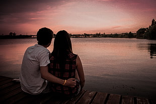 man in white shirt beside woman sitting on brown wooden dock during dusk